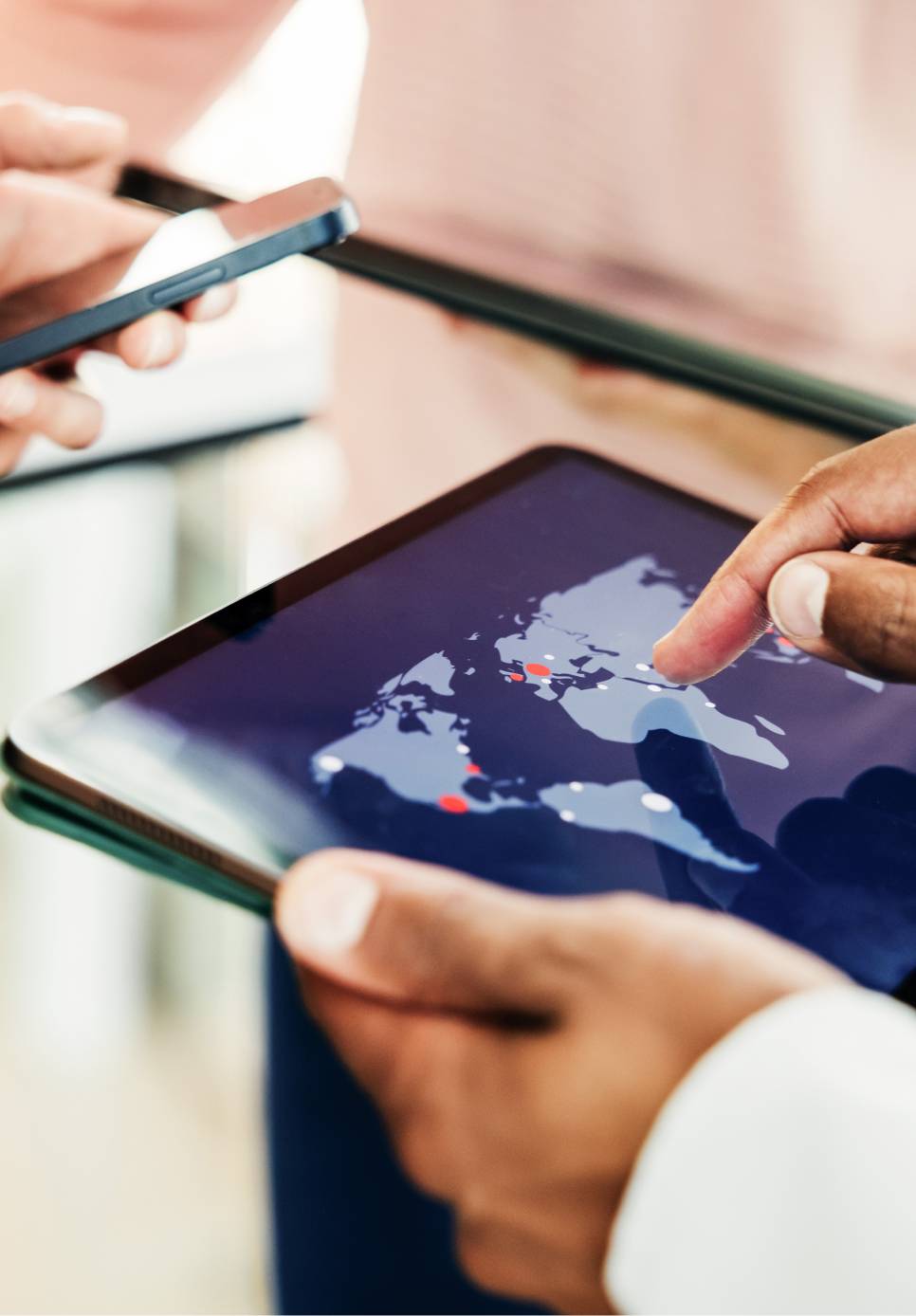 Hands holding a tablet, pointing at a map of continents with various locations, while the other hand holds a mobile phone.
