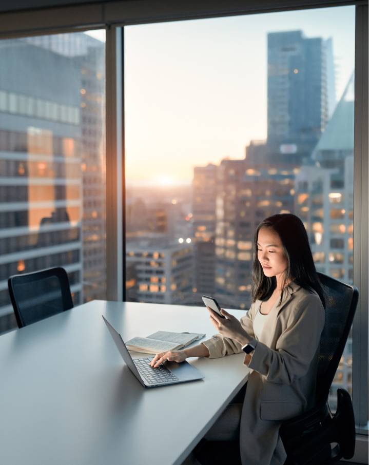 Woman in the office with a panoramic view, working on laptop and looking at mobile phone.